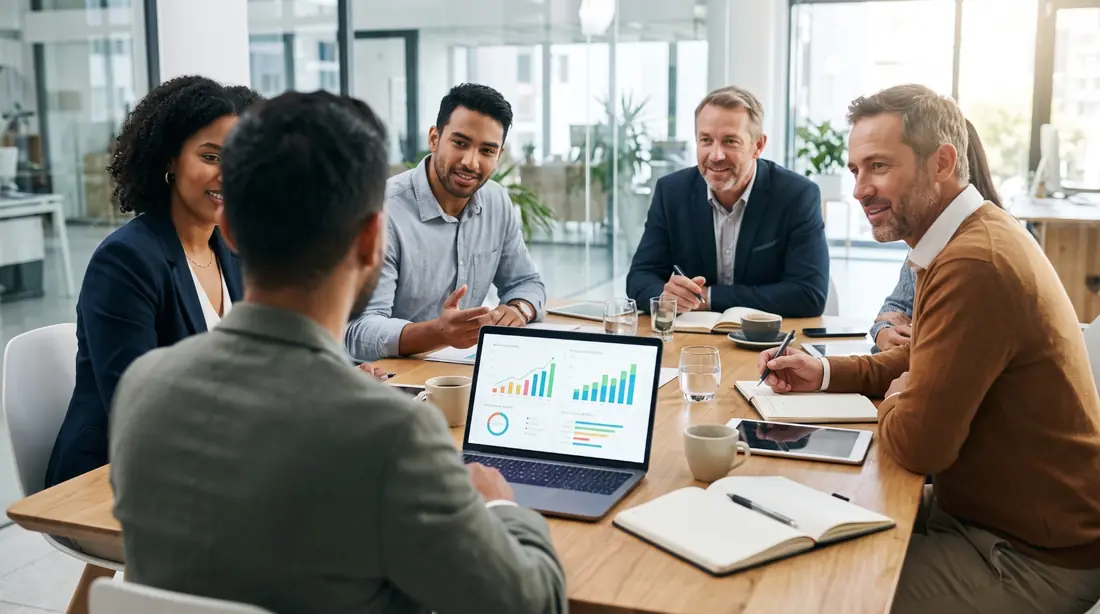 Team around a meeting table discussing performance charts on a laptop